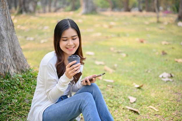 Charming Asian woman sipping coffee relaxing and sitting on grass under a tree in the park