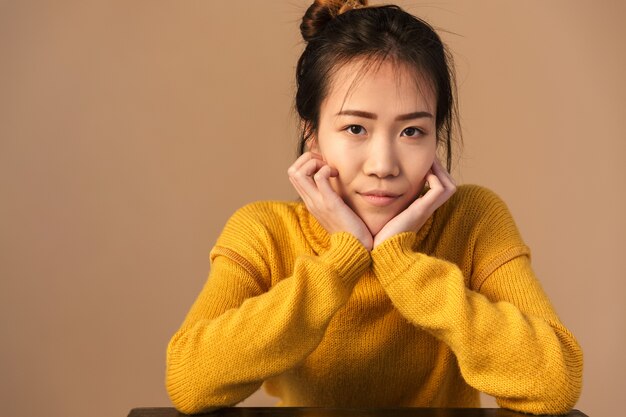 charming asian woman wearing sweater smiling while sitting in studio isolated over beige wall