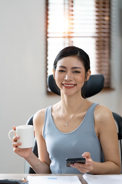 Affascinante donna asiatica con un sorriso che tiene una tazza di caffè e un telefono cellulare seduto in ufficio