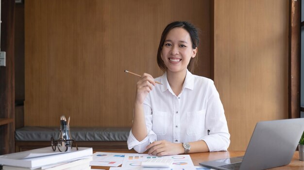 노트북을 사용하여 사무실에서 일하는 매력적인 아시아 여성 카메라를 보고