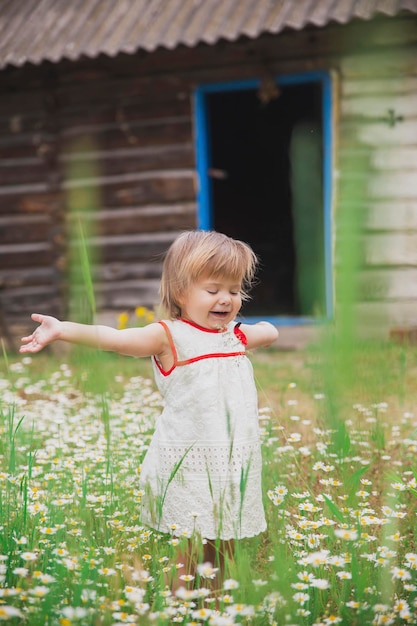 charming baby in an embroidered dress enjoys flowers near a wooden hut