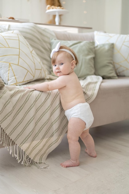 A charming baby girl in diapers is standing near the sofa at home in a bright living room with a support the child is learning to walk the first steps