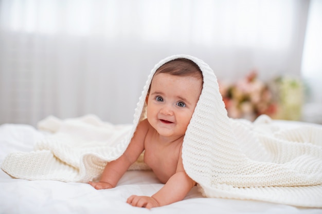 Charming baby girl is lying on the bed under a white blanket and smiling.