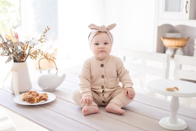 A charming baby girl in a jumpsuit made of natural fabric is sitting on the table at home in a bright kitchen the first lure