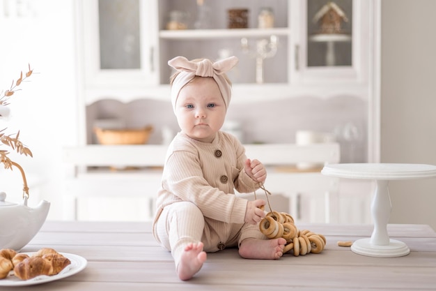 천연 직물로 만든 점프수트를 입은 매력적인 여자 아기가 밝은 부엌에서 첫 번째 미끼로 집에 있는 테이블에 앉아 있다