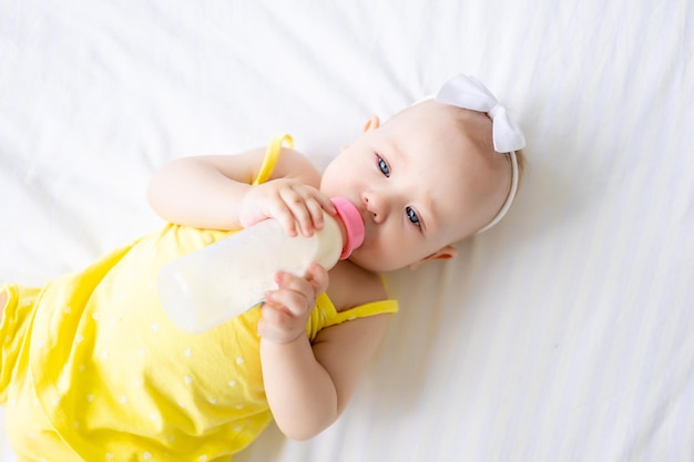 Charming baby girl in a white sunny bedroom A newborn baby is lying on the bed and sucking a bottle of milk closeup Baby food infant formula