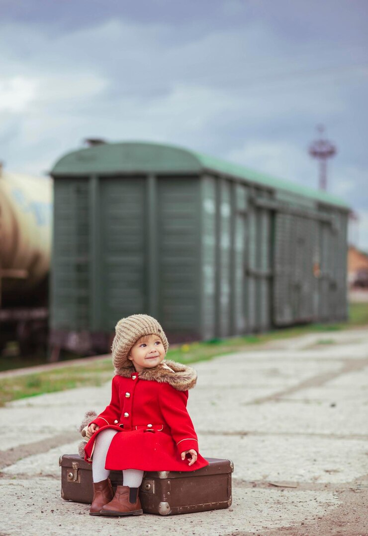 Photo charming baby in a red coat with a suitcase is waiting for a train on the platform