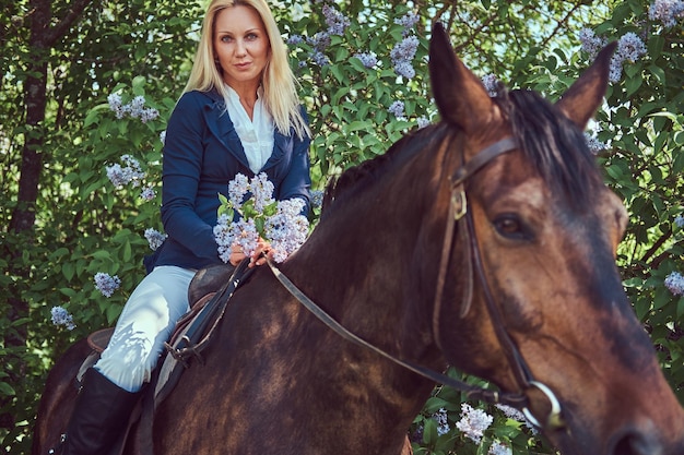 Charming beautiful blonde jockey riding a brown horse in the flower garden.