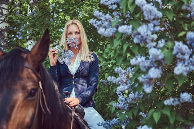 Charming beautiful blonde jockey riding a brown horse in the flower garden.