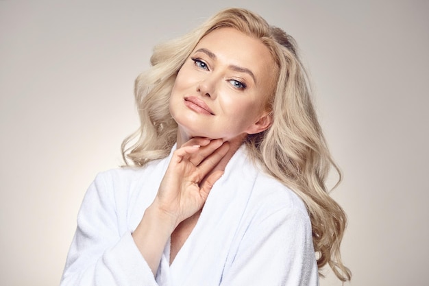 Charming beautiful looking adult blonde woman in her fifties wearing a spa robe put her hand near face having a clean and healthy skin on a beige isolated