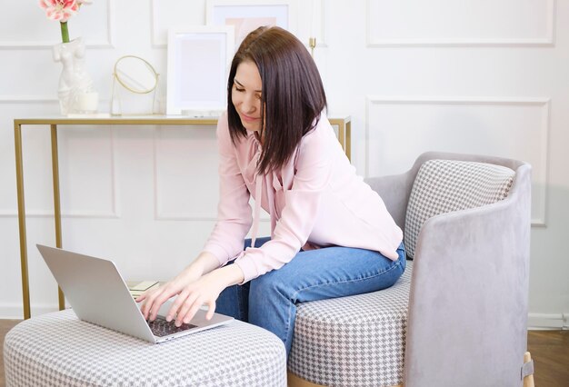 Charming beautiful young girl with curly hair sits on a blue sofa at home in front of a laptop, remote work and education, beauty and fashion.