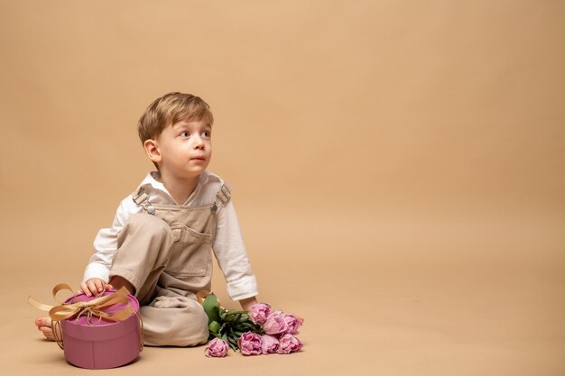 a charming blond fouryearold boy in a beige jumpsuit and a white shirt sits and holds a lilac gift