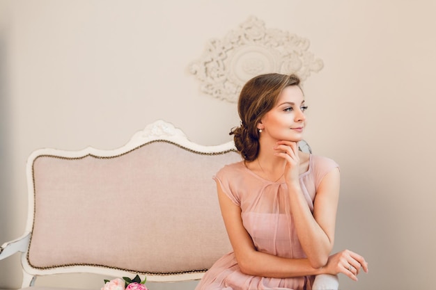 Очаровательная белокурая девушка, сидящая на софе. У нее заплетены волосы, и она держит руку возле подбородка. Она носит светлое платье персикового цвета и смотрит в окно.