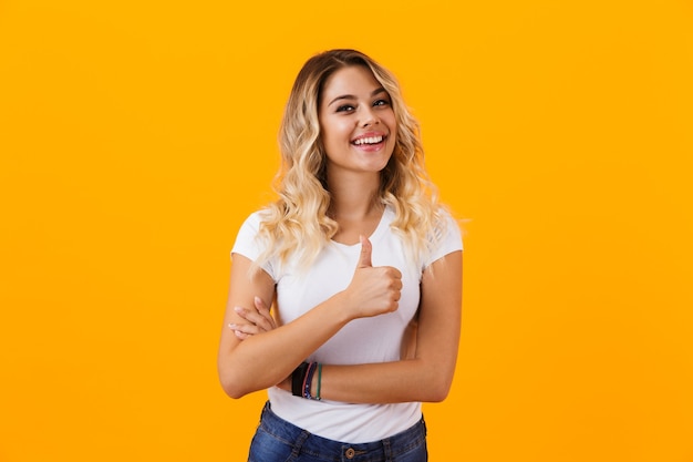 Charming blond woman in basic clothing smiling and showing thumb up, isolated over yellow wall