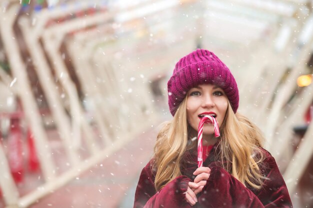 Charming blond woman wears knitted hat eating sweet candy cane at the winter street market. Empty space