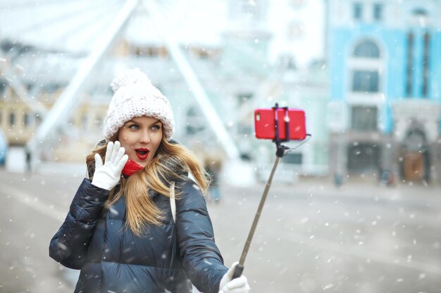 Charming blond woman with long hair taking selfie on a central city square during snowfall. empty space