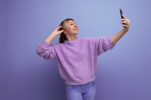 Affascinante giovane donna millenaria bionda in un look casual prende un selfie su uno smartphone su un luminoso