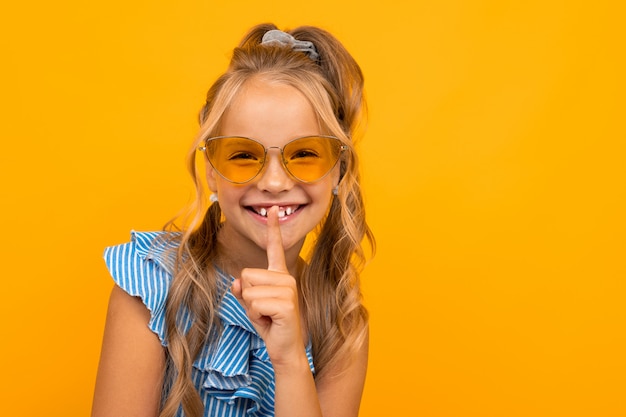 Charming blonde girl in a dress and sunglasses asks to be quieter, holds a finger at her mouth on yellow with copy space