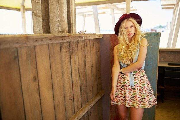 Charming blonde girl with curly hair wear burgundy hat isolated at the wooden building background