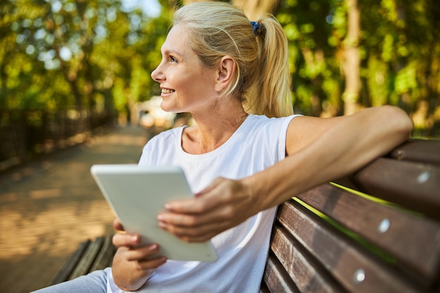 Charming blonde lady holding electronic pad PC and smiling while sitting on bench outdoors