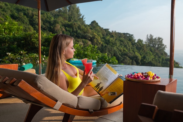 Charming blonde relaxes on a sunbed near the infinity pool and drinks a watermelon smoothie. Rest in tropical countries. Natural landscape