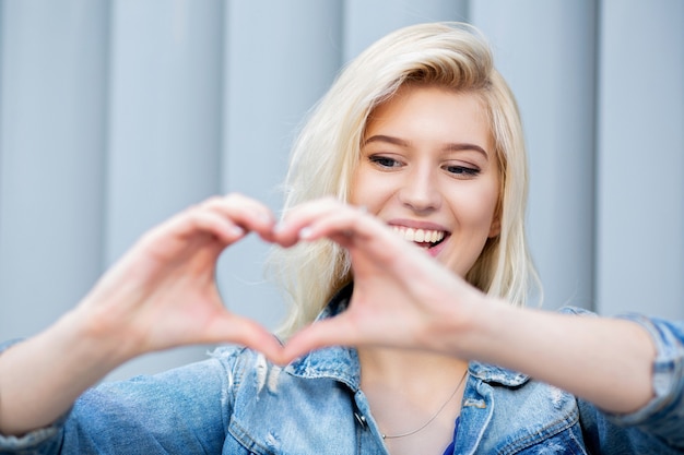 Charming blonde woman making heart sign with her fingers at the shutters background