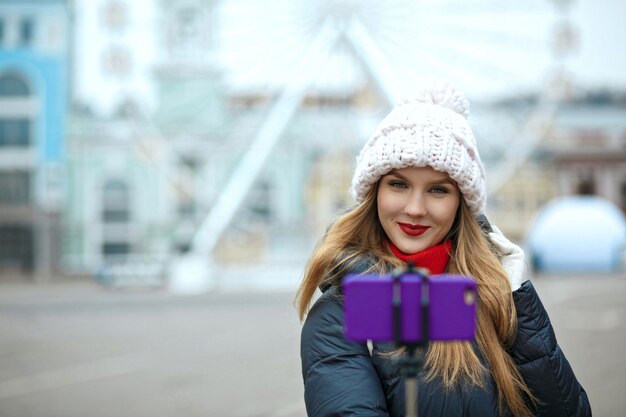 Charming blonde woman tourist wearing knitted cap, taking selfie at the street in winter. Space for text