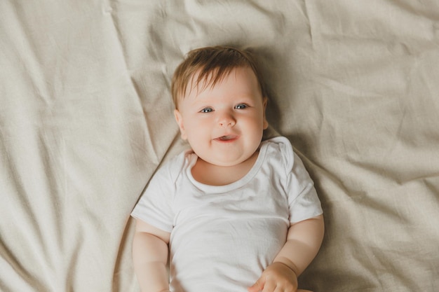 Charming blueeyed 6monthold baby lies in bed in a white bodysuit View from above