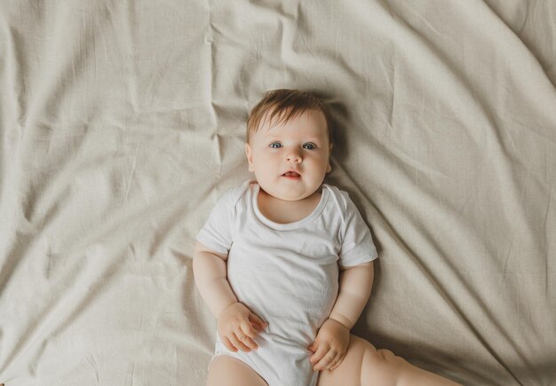 Charming blueeyed 6monthold baby lies in bed in a white bodysuit View from above