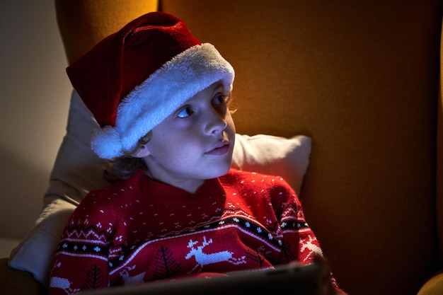 Charming boy in warm sweater and Santa hat with tablet relaxing in soft armchair while looking away in dark room with lamplight