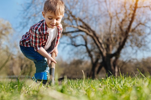 A charming boy wearing plaid shirt digging a hole in the ground to plant a tree in backyard