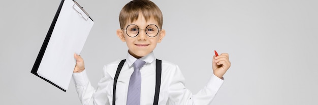 A charming boy in a white shirt, suspenders, a tie and light jeans stands. The boy holds a pen and sheets for notes