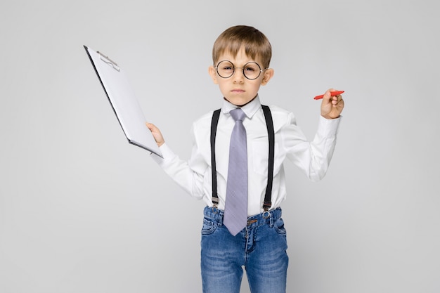 A charming boy in a white shirt, suspenders, a tie and light jeans stands on a gray background. The boy holds a pen and sheets for notes