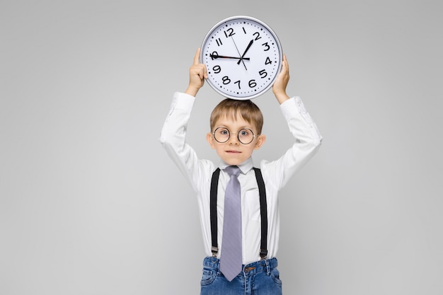 A charming boy in a white shirt, suspenders, a tie and light jeans stands on a gray . A boy holds a watch on his head