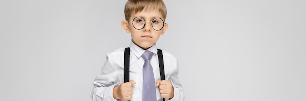 A charming boy in a white shirt, suspenders, a tie and light jeans stands on a gray wall. 