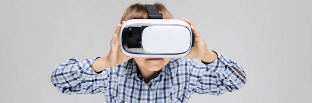 A charming boy with an inlaid shirt and light jeans stands on a gray background. The boy on his face glasses virtual reality