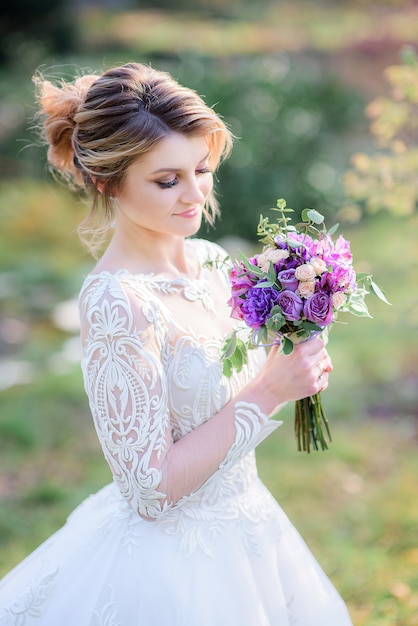 Charming bride poses with violet wedding bouquet on the green lawn