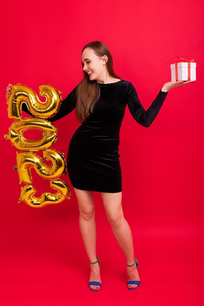 Очаровательная брюнетка в черном платье и на каблуках с подарочной коробкой держит фигурки нового 2023 года на красном фоне