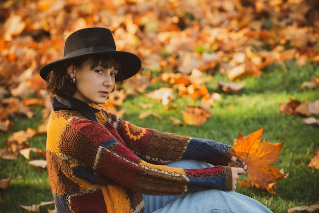 Charming brunette in hat with autumnal leaves. Autumn queen wearing cozy sweater. Autumnal forest in golden and yellow colors. Hipster woman in vintage hat walking in park.