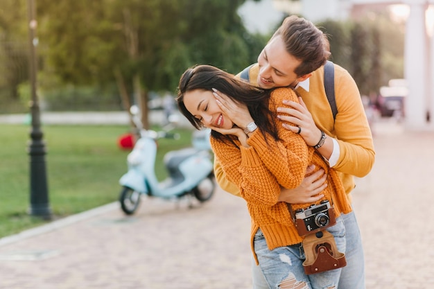 Charming brunette lady in cozy attire touching face while boyfriend embracing her on alley in park. Outdoor portrait of trendy girl with camera in orange sweater hanging out with husband on the street