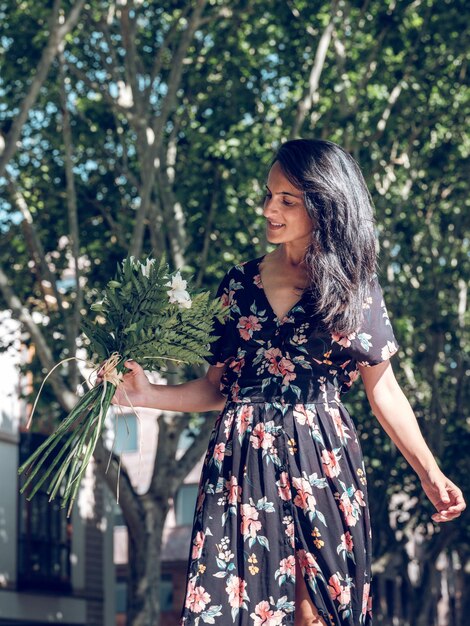 Charming brunette wearing black summer dress looking at green bouquet with white flowers while walking along city alley in sunny day