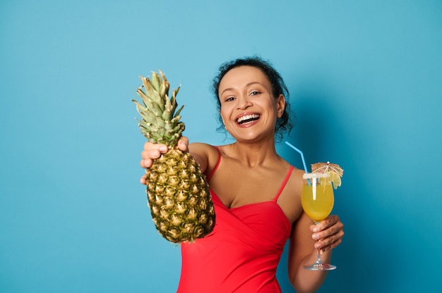 Affascinante bruna con un bel sorriso che tiene un bicchiere con cocktail e mostra un ananas alla telecamera.