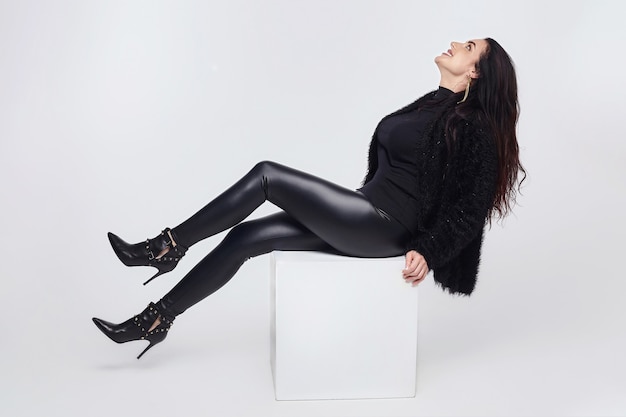 Charming brunette with long hair is sitting on a white cube. photo session in the Studio on a white background