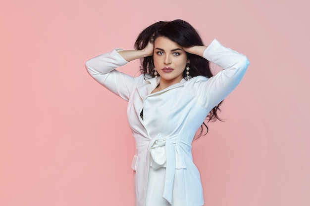 Charming brunette with long hair and a white suit. photo shoot on a pink background