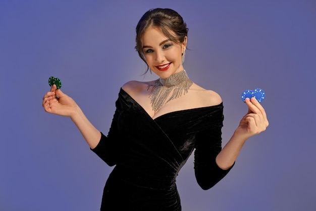 Charming brunette woman, bare shoulders, bright make-up, in black dress and shiny jewelry. Smiling, showing two blue and green chips, posing on purple background. Poker, casino. Close-up, copy space