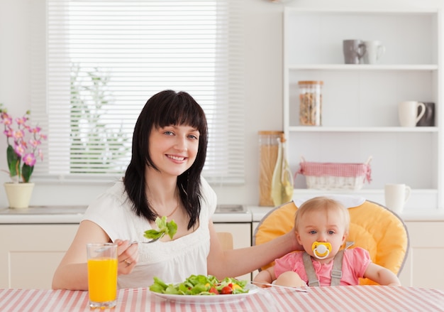 Charming brunette woman eating a salad next to her baby while sitting