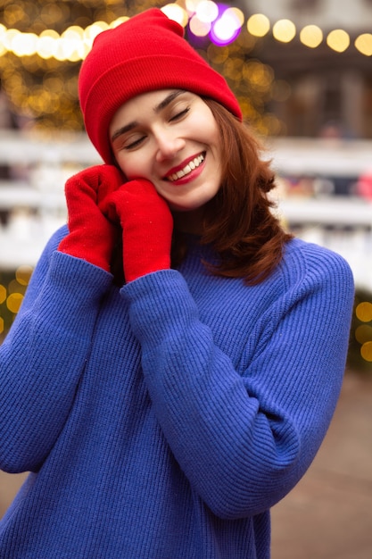 Charming brunette woman wears red cap and blue sweater walking at the Christmas fair in the central city square