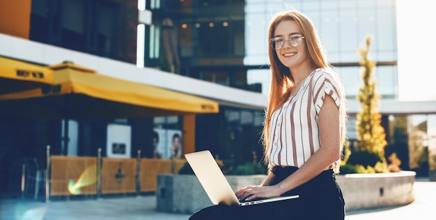 Charming businesswoman with eyeglasses and red hair posing outside with a laptop sitting on the bench