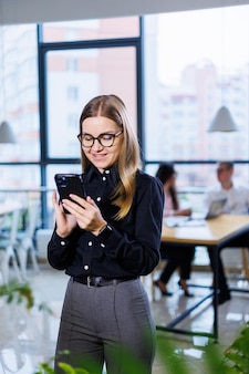 Affascinante donna d'affari con gli occhiali vestita tutti i giorni, utilizzando uno smartphone e parlando al telefono, in un ufficio moderno.