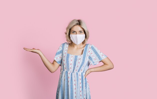 Charming caucasian blonde woman advertising something with medical mask on face on a pink studio wall with free space gesturing with palm